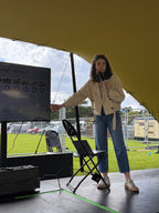 Nuala on stage with a microphone pointing at a screen, with a tent and outdoor setting in the background.